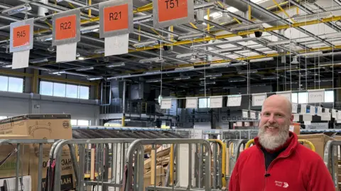 Jersey Post Chris wearing his red Jersey Post fleece jacket standing in front of trollies of parcels in the Jersey Post headquarters