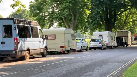 A row of caravans and vans on the side of the road with a few cars in between, they are parked next to large grassy open spaces with large trees.