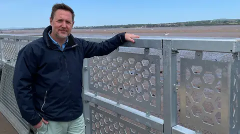 A man called Stuart Greenslade stands next to a fence on a train station platform. He is wearing a navy blue coat. He has brown trimmed hair and a trimmed grey beard. The River Exe is behind the railings.