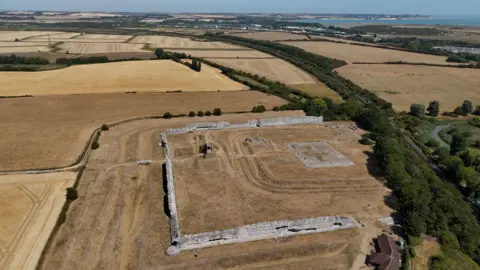 Richborough Fort, which is a square stone ruin, seen from above. The ruined walls contain yellow grass, which also surrounds the site.