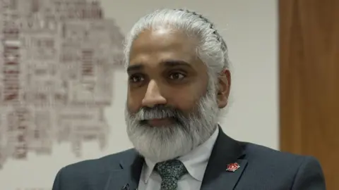 A man wearing a dark grey blazer, white shirt and dark grey patterned tie sits down looking at an angle