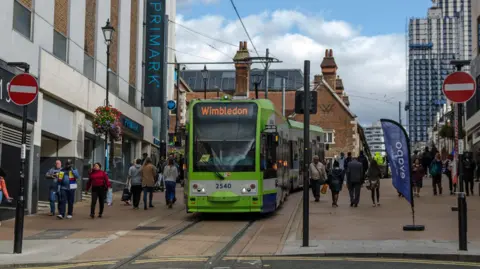 A green tram in a shopping street in Croydon. There are shoppers on either side of the tramway 