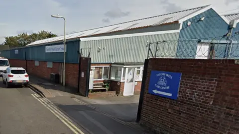 A corrugated metal warehouse at Jacobs Well Care Centre. The entrance gate is between brick walls topped with coiled, barbed wire.