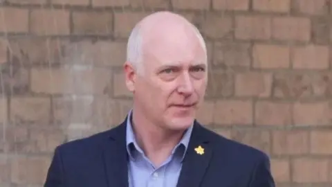 A balding man wearing a dark suit with a brick wall in the background 