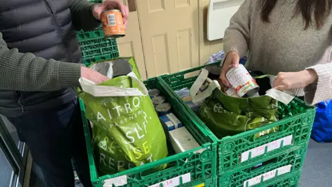 John Devine/BBC Two people pack plastic bags with various food items, tins, packets and boxes from some large green crates