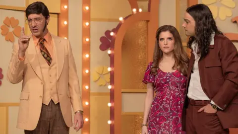 Netflix Tony Hale in a beige and brown 70s suit, Anna Kendrick in a red paisley dress and Daniel Zovatto in a brown 70s suit in the Netflix series “Woman of the Hour”.