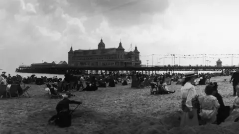 Getty Images A black and white image of what Britannia Pier looked like in Great Yarmouth in 1910. The pier stretches out across the sea, on the right side of the image, and lots of people are sat on the beach.