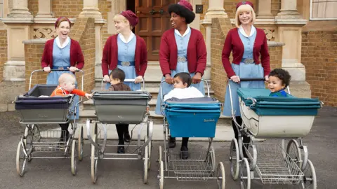BBC/Neal Street Productions/Nicky Johnston Four of the stars of Call the Midwife push prams with children in them away from a brick building. They are all women and wear blue uniforms with burgundy cardigans and burgundy hats.