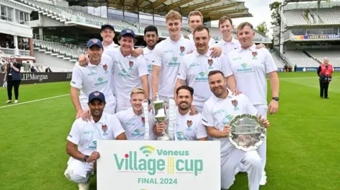 Paul Carroll Photography Cricket team with trophy in front of sign saying Voneus Village Cup final 2024. Thirteen men wearing short-sleeved white shirts are in the centre of the photograph. Four men at the front are crouching. One is holding a white sign containing the words  "Voneus Village cup FINAL 2024" in green letters. One is holding a silver cup in his right hand. One is holding a silver platter with two hands. Nine men are standing behind the four men at the front. Seats in stands can be seen in the background.