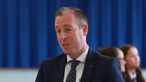 PA Media Paul Givan. He has short grey-ish hair, wearing a navy blazer, white shirt and navy tie with white polka dots. A blue curtain is behind him with two female pupils are blurred behind him.