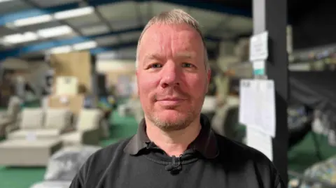 a man with short grey hair and a stubble beard looks at the camera wearing a black jumper with blurred out garden furnitre behind him