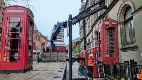 One red booth is on a lorry bed on the left while a workman helps remove another box on the right, which is being lifted by a crane. Another seven red booths are lined up behind, next to the Edwardian former post office.