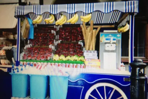 Gussy's Ice Cream A black-and-white photograph of a street stall at night. Several people stand around the counter, with one person behind the stall holding newspapers. Bottles and other items are displayed on the counter beneath a striped canopy.