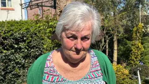 A woman with grey hair is wearing a green top. She is prominent in the photo and bushes and parts of buildings are behind her.