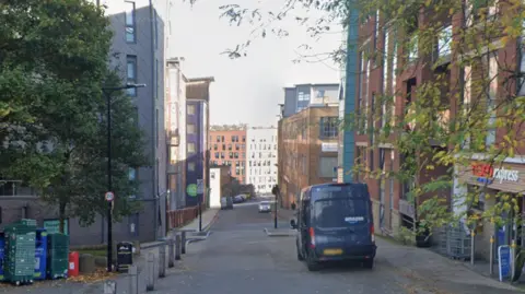 Google A narrow urban street lined with mid-rise buildings on both sides, featuring a mix of brick and modern facades. On the left, there are stacked recycling bins and a large tree with green foliage. On the right, a convenience store and a parked black van.