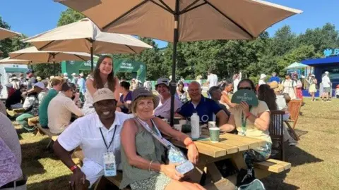 Handout A man wearing a white linen shirt and a black lanyard around his neck. He is sat next to a woman, in a green top and green dress, with a multi-coloured bag across her shoulder. Another woman is wearing a yellow shirt and holding a green fan covering half of her face. There is also a man in a blue collared shirt. They are sitting at a table with a large outdoor umbrella above them. 