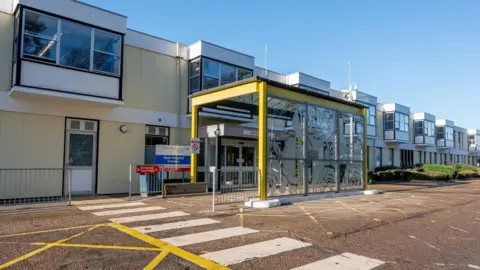 The QEH The exterior of The Queen Elizabeth Hospital. It is a modern two-storey building painted in blue and yellow. A pedestrian crossing is painted on the road in the foreground.