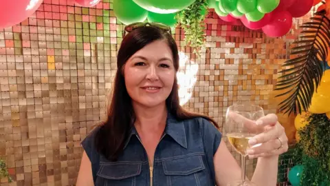 Hayley surrounded by balloons , holding a glass of wine and smiling