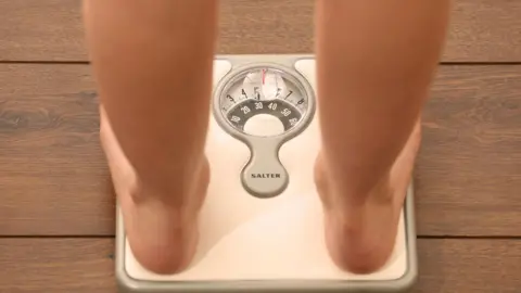 An image of someone using a set of weighing scales. Only part of their legs and feet are visible.