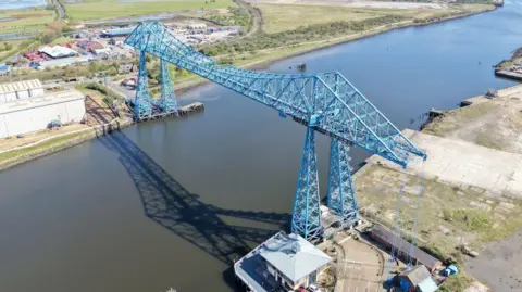 CAD Aerial A large blue metal bridge which towers over a river. There are fields on either side. 