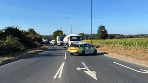 A police car is parked at an angle across a road, with traffic queued behind it. The other side of the road is empty.