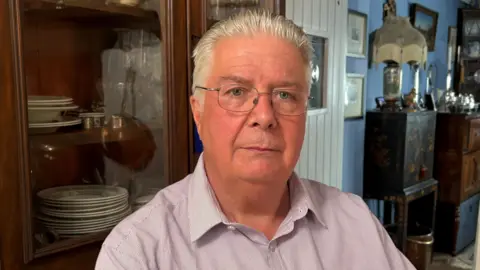 A man with grey hair, which is combed back, is sitting in a chair. He is wearing glasses and a pink, open collared shirt with pinstripes. Behind him is a collection of antique furniture and household items including a lamp and photographs. There is also a white cottage style door separating two rooms.