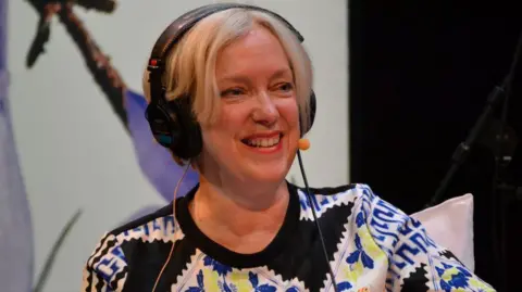 BBC Janice Forsyth mengenakan headset radio dan mikrofon. Dia memiliki rambut pirang pendek dan atasan dengan pola yang mencolok.