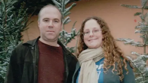 Rob Webb A man with brown hair wearing a maroon jumper and black denim jacket stands next to a woman with brown long curly hair wearing a blue shirt and cream scarf. They are smiling and stood in front of some Christmas trees with snow on the branches.