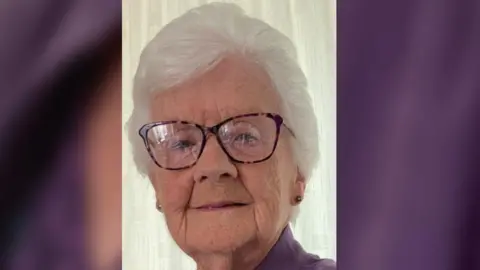 A woman in her late 80s smiles softly. She has short white hair, is wearing pattern rimmed glasses, flower stud earrings and a purple blouse. There is a white curtain behind her.