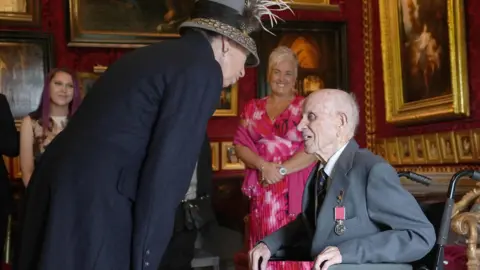PA Media The Princess Royal wearing a black coat and grey hat inside a room in Hillsborough Castle. She has a bag over her shoulder. She is speaking to Norman Irwin, who is in a grey suit and using a wheelchair. He has a medal in his pocket. There are gold-framed photos on the walls and a large fireplace. Four people are standing round the edge of the room, smiling.