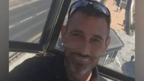 A smiling Adam Lucas has short hair and grey beard and has sunglasses balanced on his head.