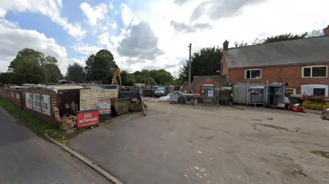 The entrance to an industrial site on Little Glen Road where the fire has broken out. A brick wall is alongside the tarmac entrance which leads part a brick building with cars and industrial equipment visible in the distance.