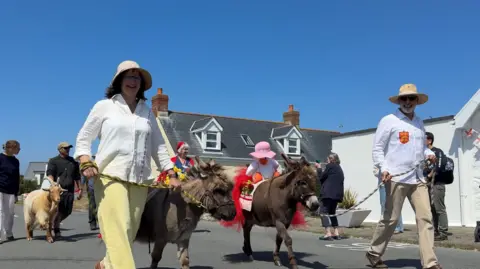 A picture of people walking with donkeys and goats along the streets. They are wearing white shirts with the donkeys on a lead.