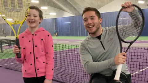 Helga Foyster A standing girl and a man in a wheelchair are side by side, smiling at the camera. The girl is wearing a pink zip up top with black spots and black trousers and is holding up a yellow tennis racket. The man is wearing a grey Ellesse zip-up top and black trousers and holding a black tennis racket. Behind them is an indoor tennis court with purple and green flooring 
