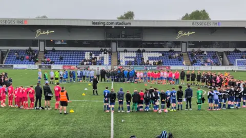 Football 4 Macclesfield A gathering of people in football kits of various colours standing around a centre circle, looking towards the centre, with a stand with blue seats in the background