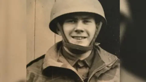 Handout A sepia tone photo of Danny Gibbon at 17 years old in the army- wearing a camouflage jacket and rounded helmet with a chin strap