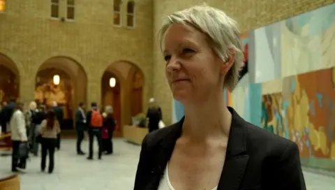 Ingrid Fiskaa in a large hall. She has short blonde hair and is wearing a white shirt and black jacket