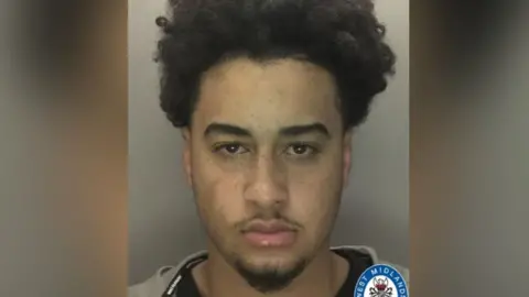 A mugshot of a man who has a short beard and thin moustache. He has brown eyes and an afro. He is staring into the camera and is wearing a grey hoodie over a black top. 