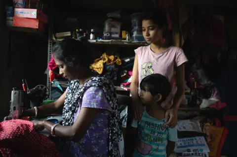 Swastik Pal Soma Das uses the little money she gets from benefits to stitch clothes and sell from her joint family home in Bishnupur. She's seen here with her daughter in their village home. Case study of West Bengal benefits.