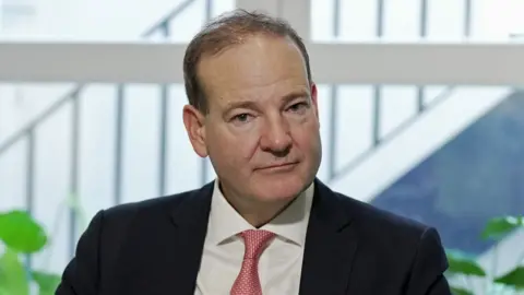 Headshot of David Garofalo who has a serious expression on his face and short brown hair. He is wearing a black suit jacket, white shirt and pink tie. Behind him is a window with plants in the foreground. 