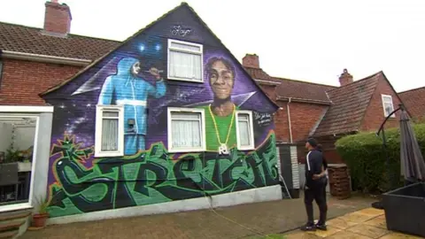 Vincent and Sandra Nembhard embrace eachother as they look up to a mural on their back garden wal. The mural has a purple background and has two paintings of Takayo. The painting on the left is of him wearing a blue tracksuit and rapping into a microphone. The painting on the right is a portrait of him wearing a green t-shirt and a gold chain around his neck. The word 'stretch' is written in black and green graffiti font at the bottom wall.