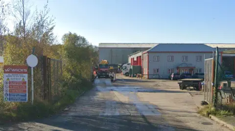 Google A Google Streetview screenshot of the entrance of Wicks, on Scarth Road, Sowerby Woods Business Park in Barrow. There is a sign with the name of the business and some safety instructions on the left of the gate, which is open and shows a large concreted industrial estate yard, with some buildings and parked vehicles.