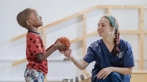 Mercy Ships A small child is practising standing while holding a toy ball, supported by a therapist in blue scrubs. They are in a rehab space with wooden steps used for therapy exercises.