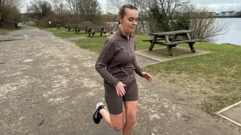 A woman in brown running top and shorts jogs along a footpath beside a lake.