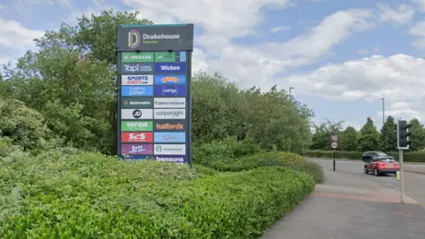The entrance area to Drakehouse Retail Park in Sheffield. A large retail park directory sign stands beside the roadside. It displays the Drakehouse Retail Park name at the top and lists a number of businesses located on the site. The sign is surrounded by hedges, greenery and trees. To the right is a road junction with traffic lights, a road sign and two cars, one red, the other black.