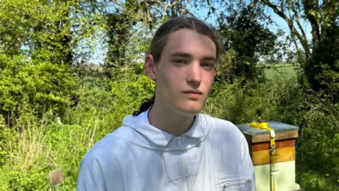 Charlie Stubbs / BBC There is a young boy in a white beekeeper uniform. He has long brown hair, tied back in a ponytail. Over his shoulder, he has a beehive with a yellow strap over the top. In the background, there is green trees, bushes and shrubbery.
