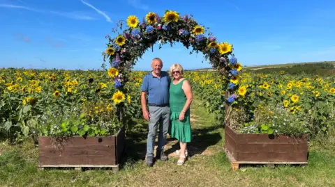 PA Real Life Hugh and Rachel are standing next to each other under an arch made of sunflowers. Behind them is a field of sunflowers.