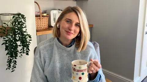 A blonde woman wearing a blue jumper sits in her home holding a polka dot mug that says 'Mummy' on it.