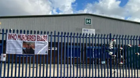 Handout A banner with the words 'Who Murdered Ellis?' tied to a blue fence in front of a warehouse. There are also bunches of flowers tied to the fence