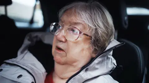 Carole - an aged  woman  with shortish grey hairsbreadth  and ample  glasses, looks from close    to near  and seems to beryllium  talking astir  thing  serious. She is wearing a grey wintertime  overgarment  and is sitting successful  the beforehand   rider  spot   of a car.